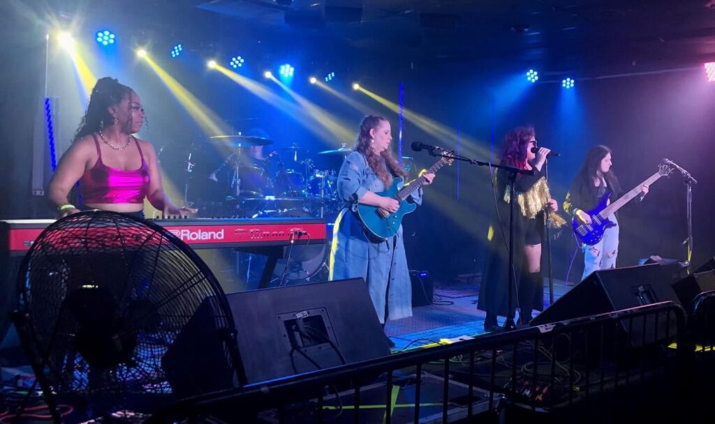 Women on stage playing keyboard, guitar, drums, bass with their singer.